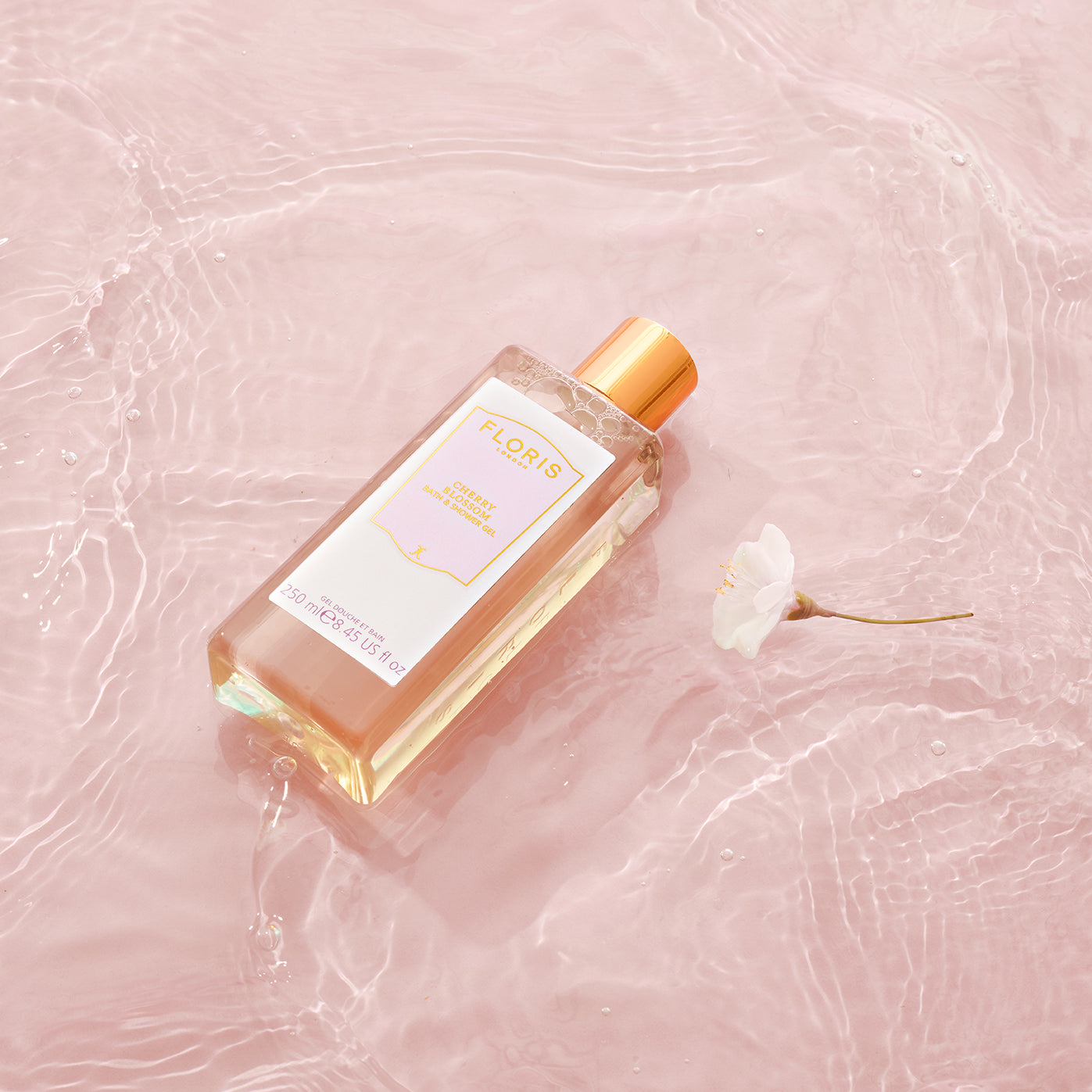 A Floris London UK Cherry Blossom Bath & Shower Gel bottle with a gold cap rests on rippled pink water, adorned with a flower.