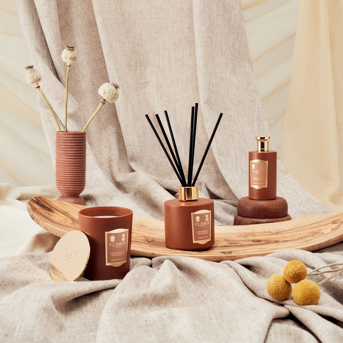 The Cinnamon & Tangerine reed diffuser from Floris London in a sleek glass vessel rests on a wooden tray, surrounded by beige fabric.