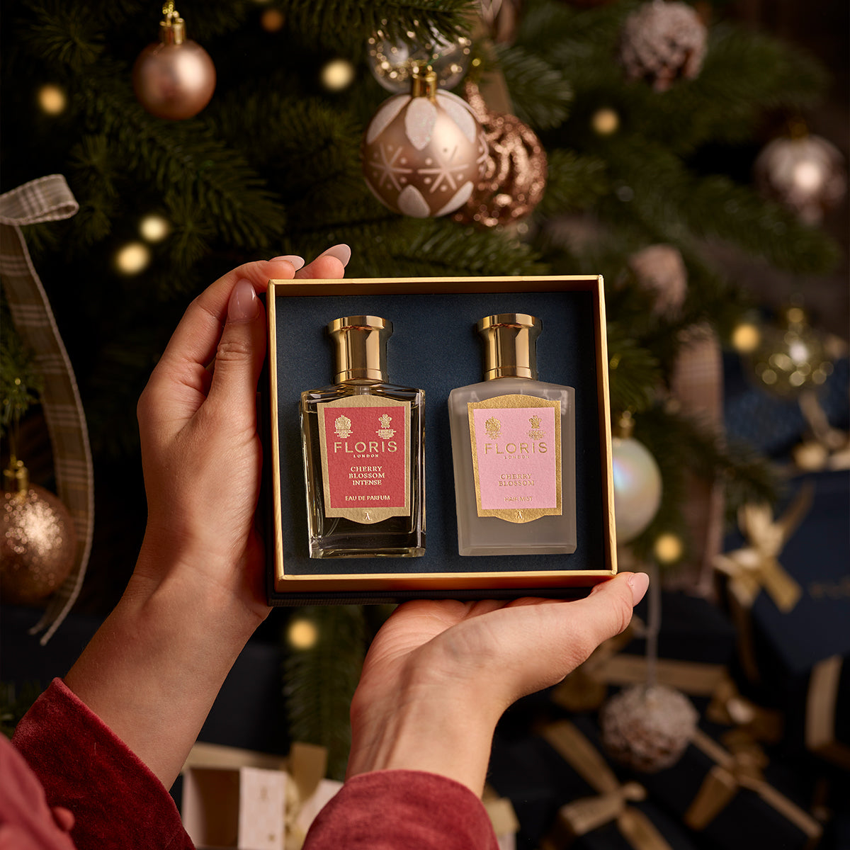 Hands holding a boxed Cherry Blossom Duet from Floris London, featuring two exquisite perfumed products, in front of a Christmas tree adorned with ornaments and lights. This luxurious gift set is perfect for the holiday season.