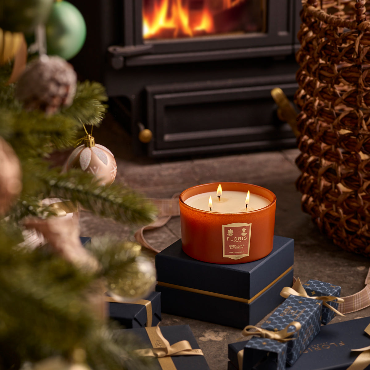 A limited-edition Cinnamon & Tangerine Deluxe Three Wick Candle by Floris London sits atop a stack of gifts near a decorated Christmas tree, with a fireplace glowing warmly in the background.