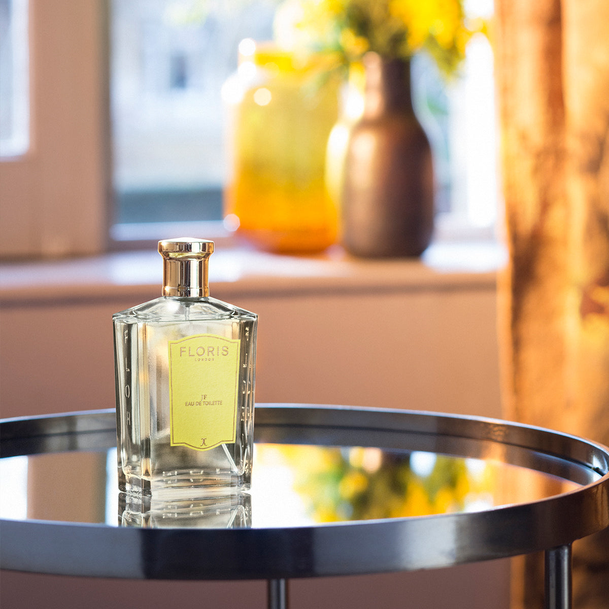 A bottle of Floris London JF - Eau de Toilette, a citrus woody scent, sits on a glass table with vases and yellow flowers blurred in the background.