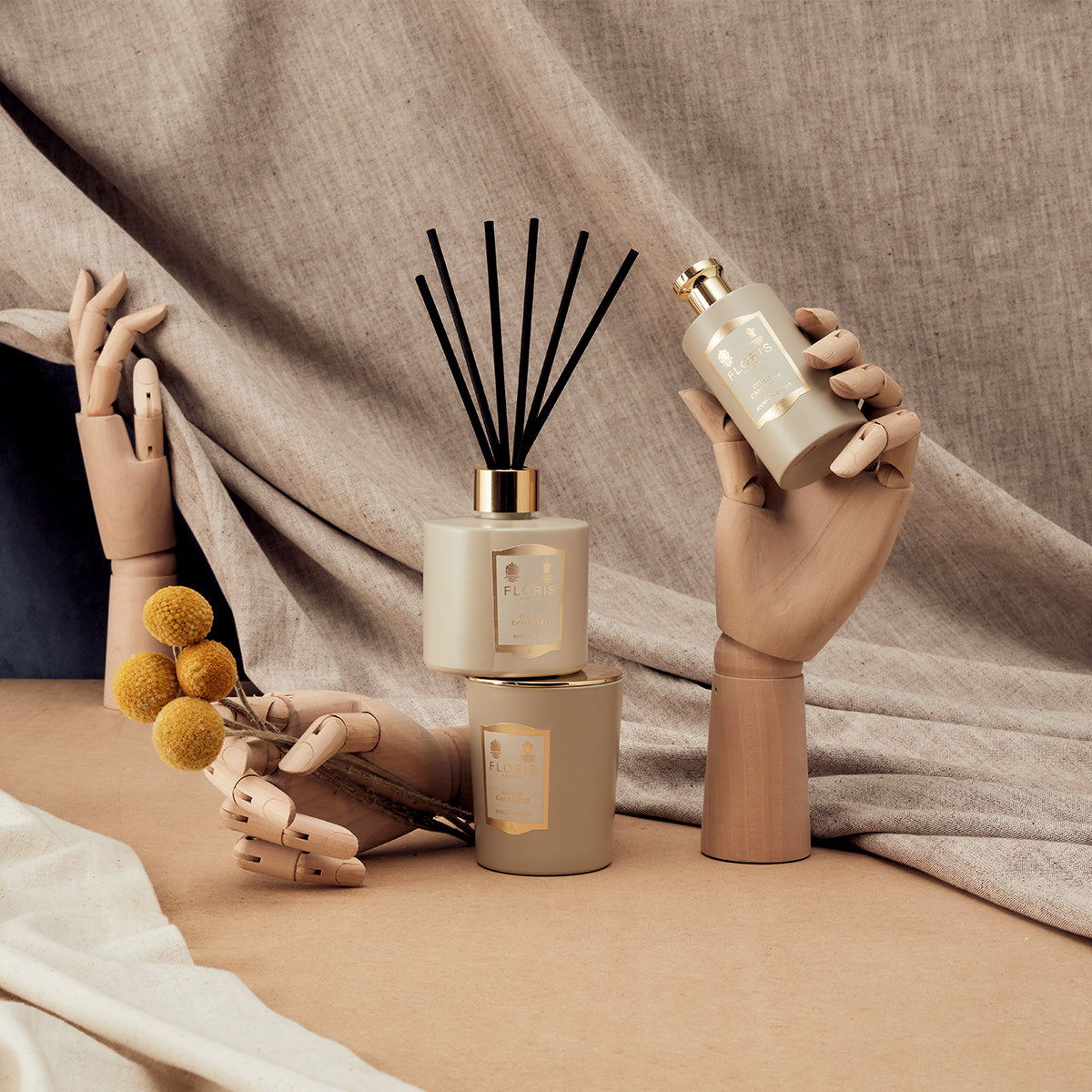 Wooden hands display Oud & Cashmere by Floris London, with candles, a diffuser, and dried flowers on a beige fabric background.