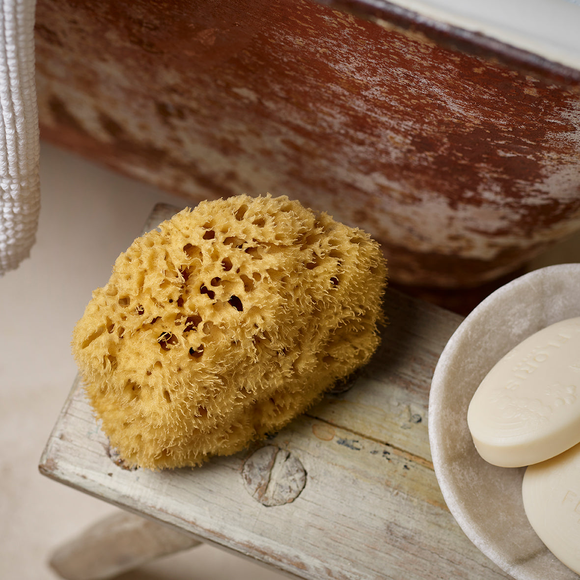 A Floris London Natural Sea Sponge and two bars of soap on a wooden stool next to a rustic bathtub promise to gently exfoliate skin.