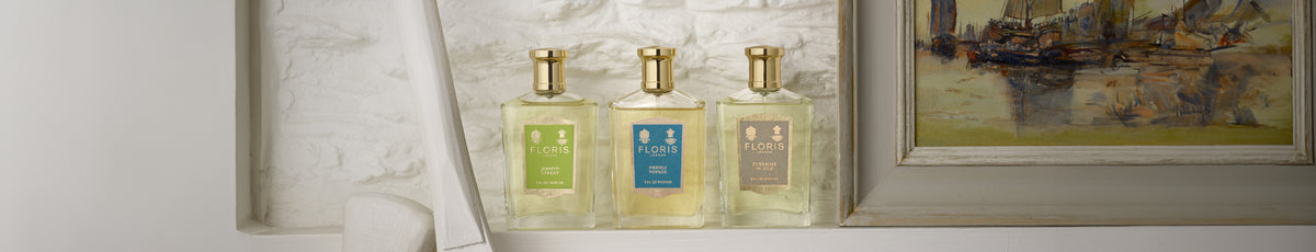 Three perfume bottles with colorful labels are displayed next to a framed painting on a white textured shelf.