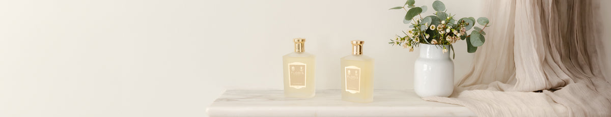 Two elegant bottles on a marble surface beside a vase with greenery and beige fabric backdrop.