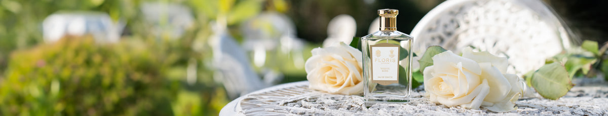 Perfume bottle and white roses on a lace table outdoors with greenery in the background.