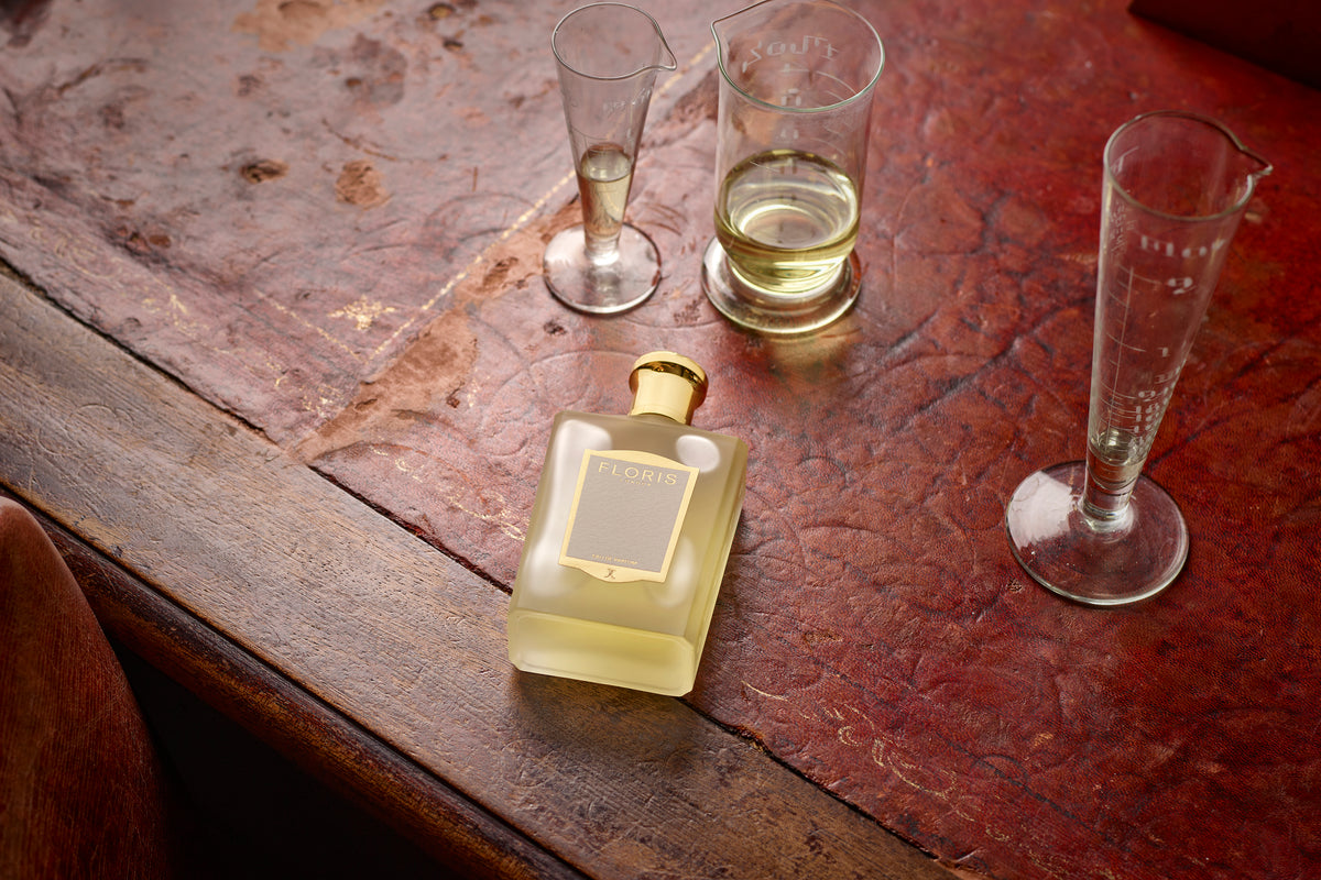 A rectangular Floris London Fragrance - Customisation Replenishment 100ml bottle with a gold cap sits on a leather table with three laboratory beakers.