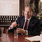 A smiling man in a suit mixes ingredients for a bespoke 100ml Fragrance Customisation by Floris London at a table with glassware and a notebook.