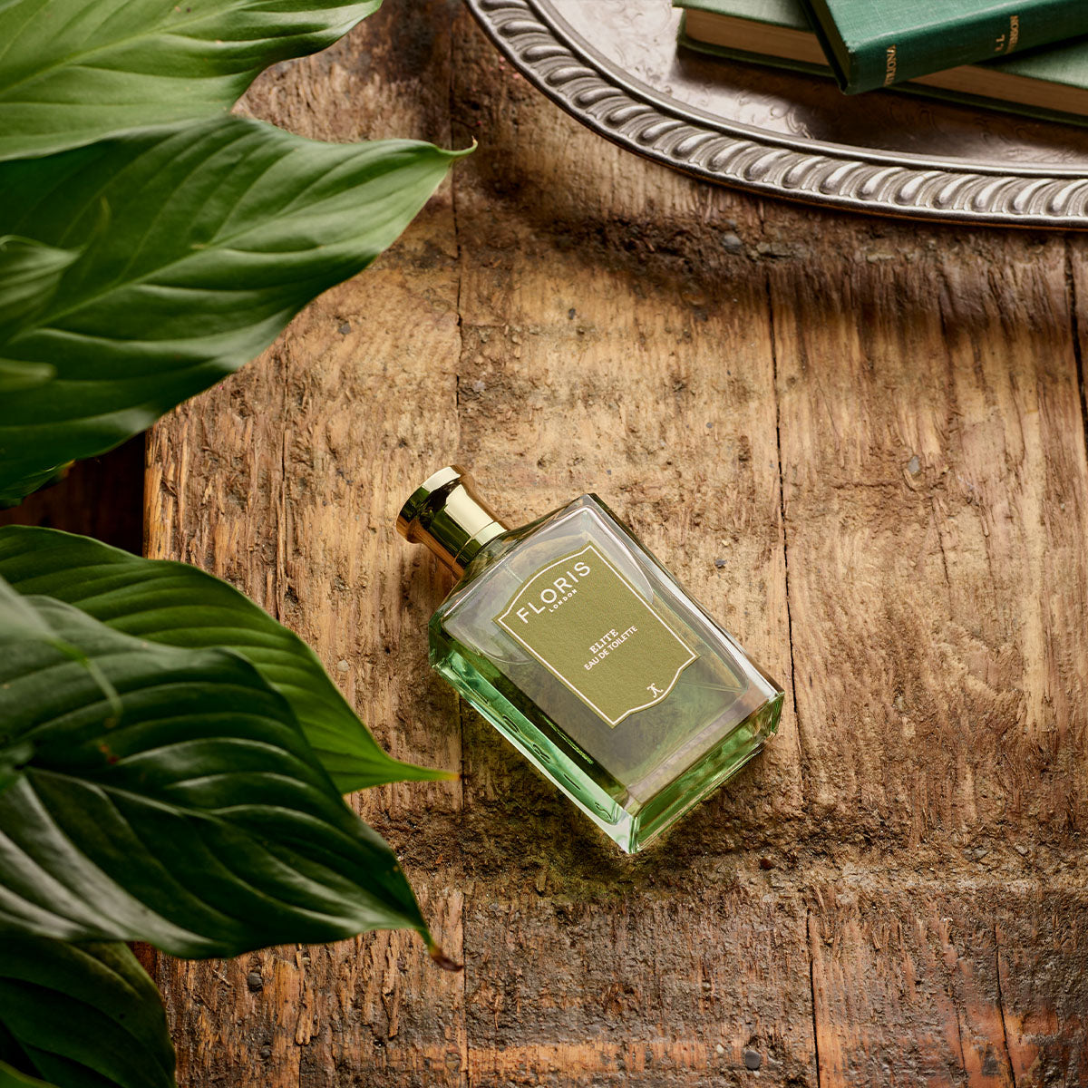 A bottle of Floris London Elite - Eau de Toilette rests on rustic wood with green leaves and a silver tray—capturing a timeless British scent moment.