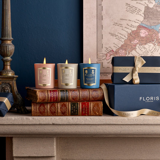 Three beautifully scented candles from the Miniature Candle Collection by Floris London sit on antique books near a decorative map, beside gift-wrapped boxes with ribbons on a mantel.