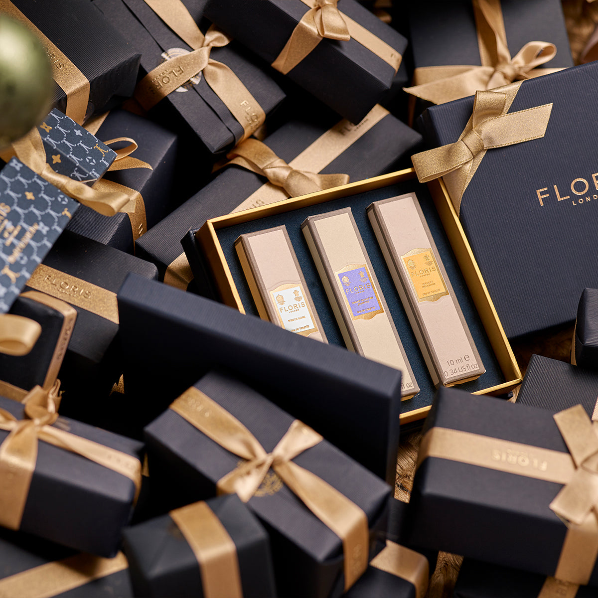 Gift boxes adorned with gold ribbons encircle a central box featuring the Floris London Perfumer's Trilogy: For Her in a luxurious setting, ideal for the season of celebration. Inside, explore exquisite floral fragrances designed to add a touch of elegance to any occasion.