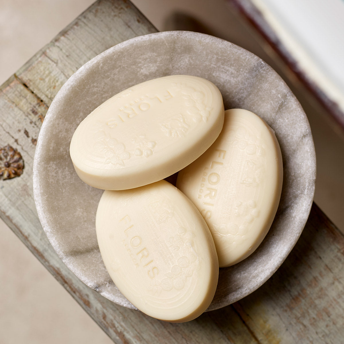 Three Soap Collection | For Her oval soaps by Floris London, with embossed floral elegance, sit in a round gray dish on a weathered wooden surface.