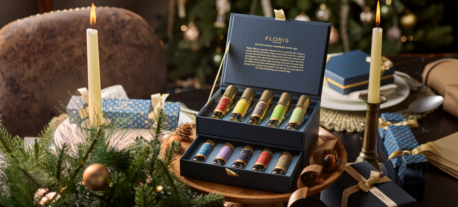 A box of assorted perfumes from The Perfumer's Collection by Floris London, with festive decor in the background, featuring a lit candle and wrapped gifts, makes for the perfect gift for any fragrance connoisseur. 