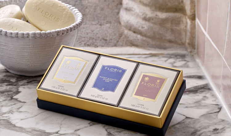 A boxed Soap Collection For Her from Floris London, featuring three exquisite soap products. The gift is in a marble bathroom with pink tiles, and next to a bowl of unwrapped soaps. This luxurious gift set is perfect for the holiday season.