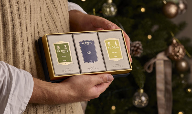 Hands holding a boxed Soap Collection for Him from Floris London, featuring three exquisite soap products, in front of a Christmas tree adorned with ornaments and lights. This luxurious gift set is perfect for the holiday season.