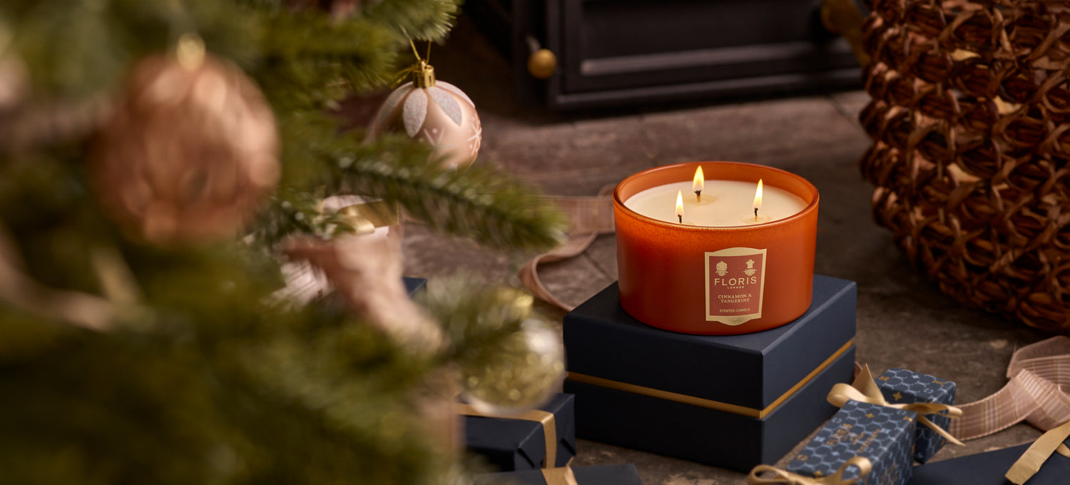 A limited-edition Cinnamon & Tangerine Deluxe Three Wick Candle by Floris London sits atop a stack of gifts near a decorated Christmas tree, with a fireplace glowing warmly in the background.