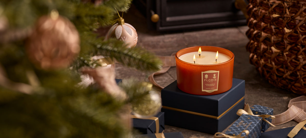 A limited-edition Cinnamon & Tangerine Deluxe Three Wick Candle by Floris London sits atop a stack of gifts near a decorated Christmas tree, with a fireplace glowing warmly in the background.