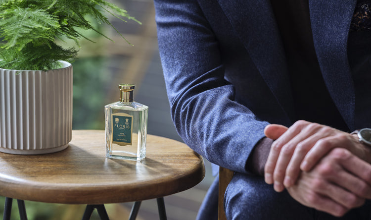 A man sat next to a small wooden table with a plan and a bottle of Floris London Vert Fougere Eau de Parfum bottle
